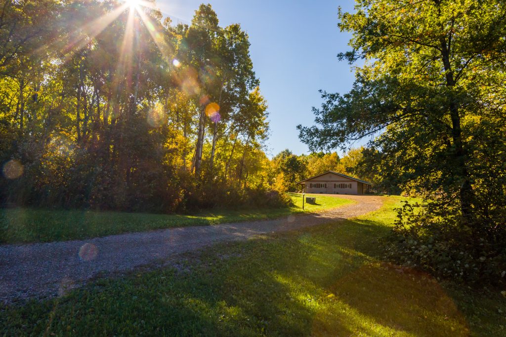 Photo of sun shining through the trees and gravel drive leading up to Diane's House at Nature's Edge Therapy Center in Rice Lake, WI.