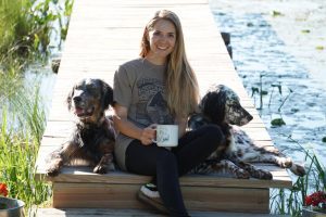 Photo of girl with long blonde hair wearing a grey tshirt and black pants and holding a coffee mug. She is sitting on a dock and has two black dogs on either side of her. 